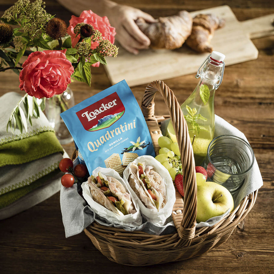 Quadratini Vanilla, Creme-Filled Wafer Cookies, 8.82 oz. Bag in a basket with sandwiches, apple, strawberries, grapes, tomatoes, soda and glass with rose next to it.