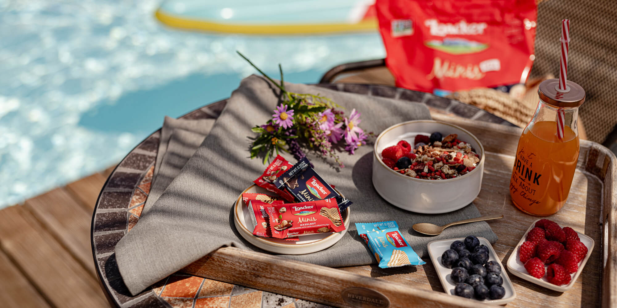Outdoor setting with Loacker Minis, snacks, berries, and drinks by a poolside.