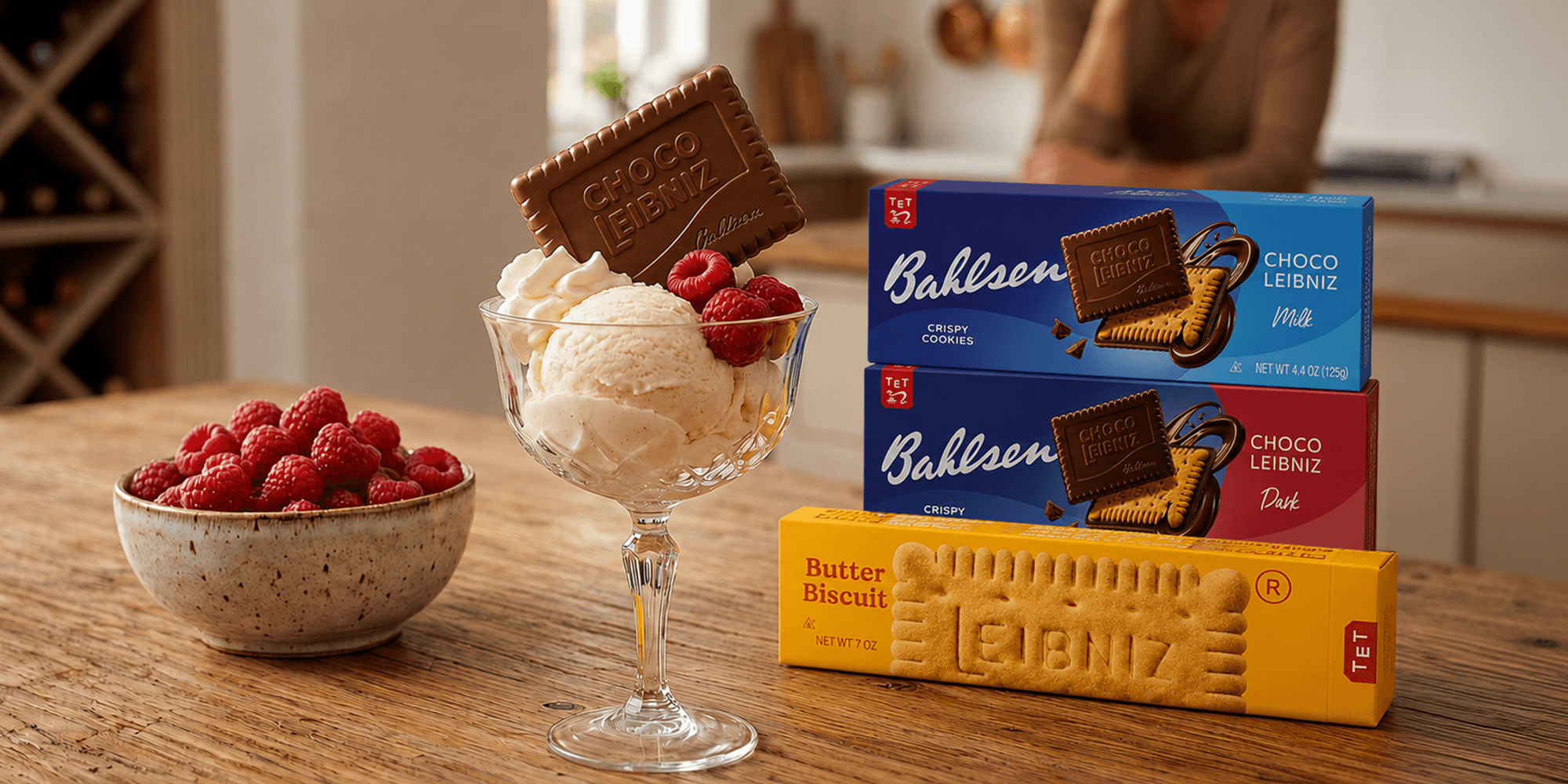 A glass dessert dish with vanilla ice cream topped with raspberries and a Choco Leibniz biscuit. Nearby, a bowl of fresh raspberries and stacked biscuit boxes.