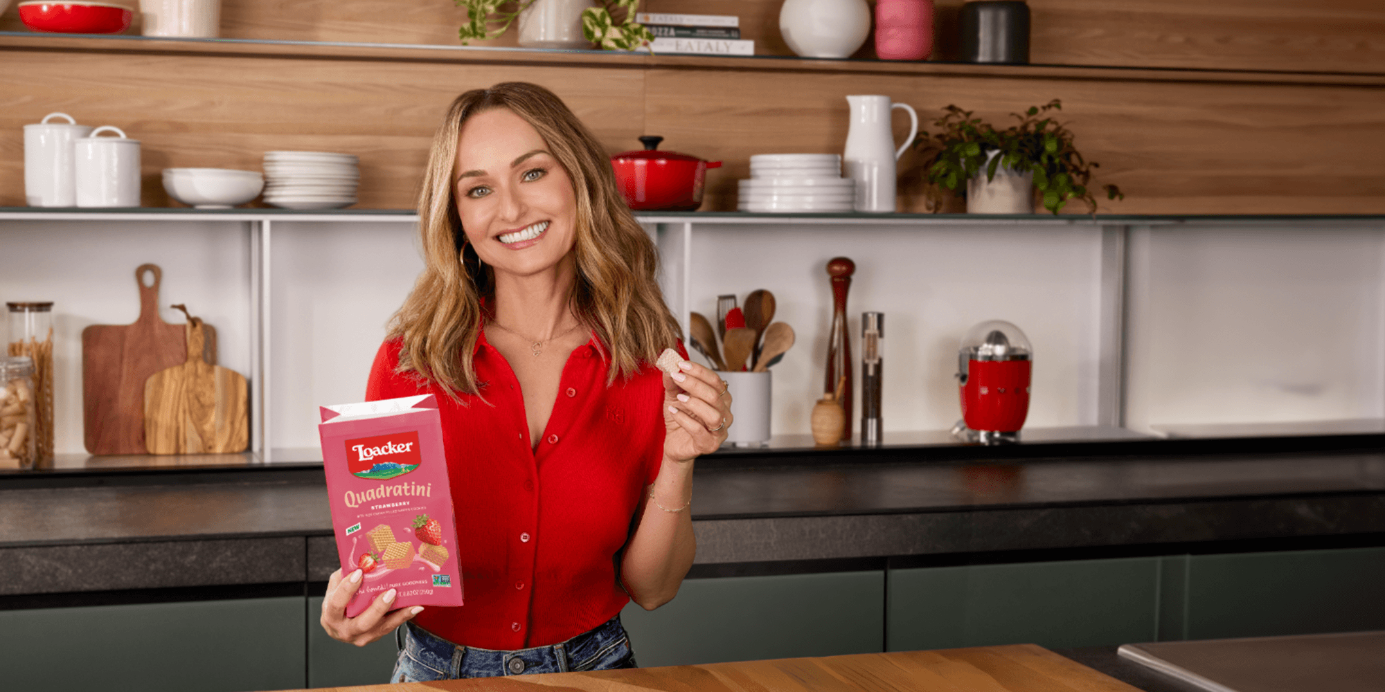 A woman in a red shirt smiles brightly in a kitchen, holding a package of Loacker Quadratini Strawberry wafers and a single wafer, conveying a cheerful mood.