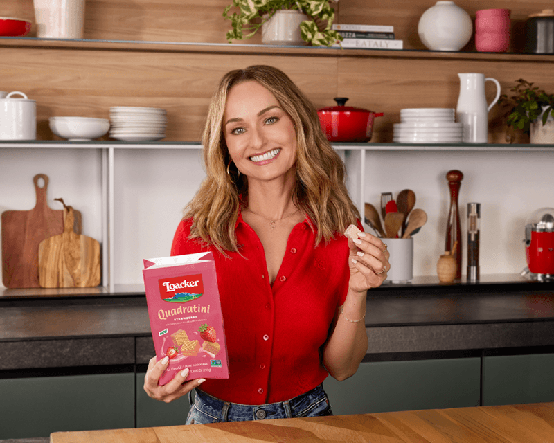 A woman in a red shirt smiles brightly in a kitchen, holding a package of Loacker Quadratini Strawverry wafers and a single wafer, conveying a cheerful mood.