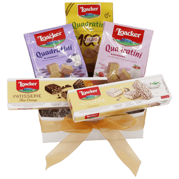 Gift box filled with Loacker products, including Quadratini and Patisserie wafers. Packages display flavors like blueberry and coconut, tied with a gold ribbon.