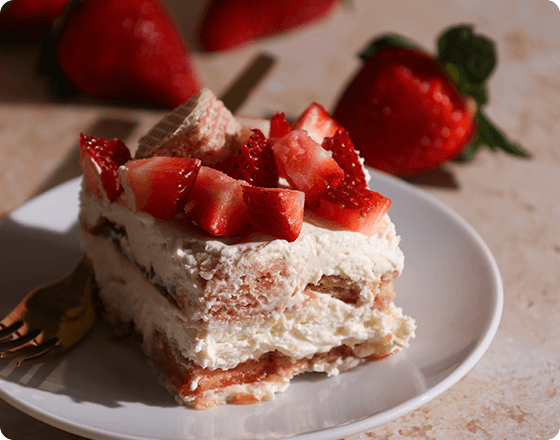 A slice of creamy strawberry cake topped with fresh chopped strawberries on a white plate. In the background are whole strawberries, creating a delightful and fresh atmosphere.