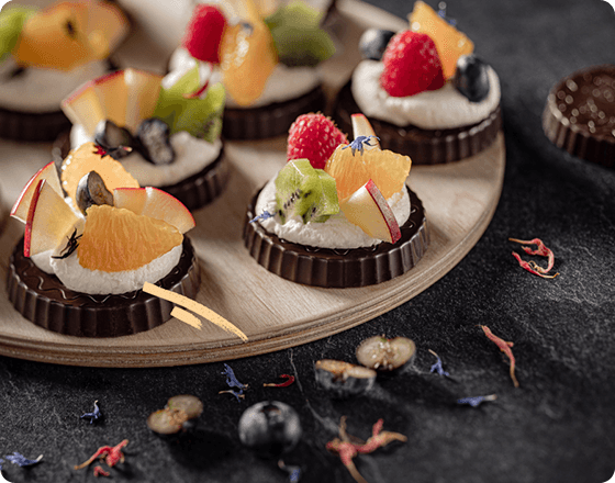 A wooden tray holds artfully arranged chocolate tarts topped with whipped cream and vibrant fruit slices like kiwi, raspberry, and orange on a dark background.