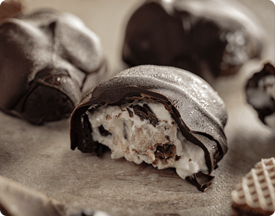 Close-up of a chocolate-covered dessert with a creamy, textured filling, possibly with nuts or nougat, on parchment paper. The scene feels indulgent and rich.
