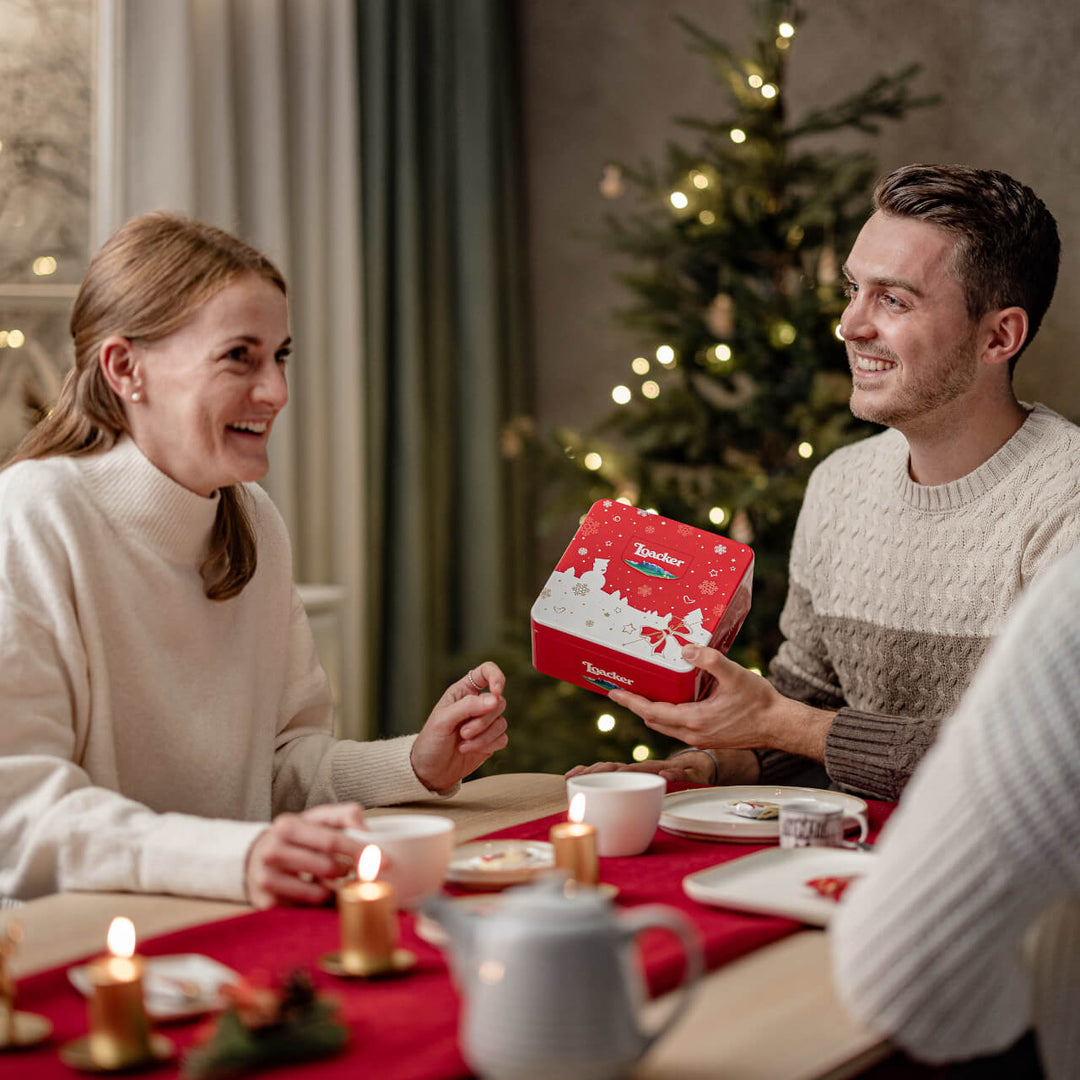 Man presenting a Tunnock's Tea Cake to a woman at a festive table with Christmas decorations.