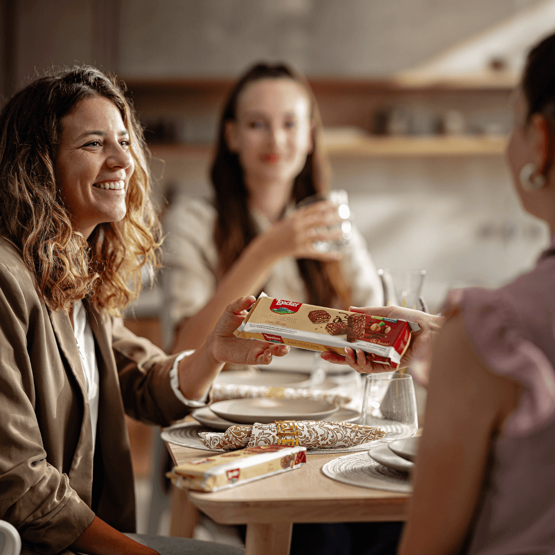 Woman holding a box of Patisserie Crème Noisette, Chocolate Coated Specialty, 3.53 oz. packaging at a table with friends in a kitchen setting