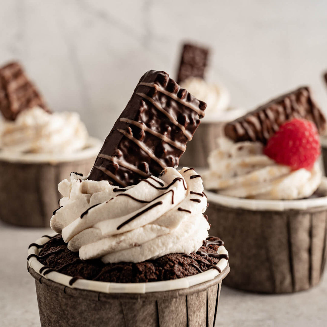 Chocolate cupcake with whipped cream and Patisserie Dark Hazelnut, Chocolate Coated Specialty on top, with blurred cupcakes in the background.