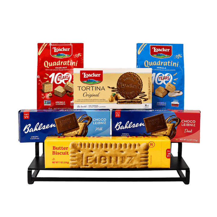 A multi-item assortment of Lóacker and Bahlsen cookies displayed on a black metal stand against a white background. The assortment includes Lóacker Quadratini in red and blue boxes, Lóacker Tortina Original in a cream box, and Bahlsen Choco Leñiz in blue and dark blue boxes, along with a yellow Butter Biscuit Biscuit.